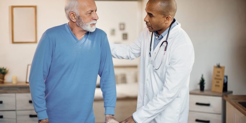 Black doctor talking to senior patient while helping him to walk with mobility walker at nursing home.