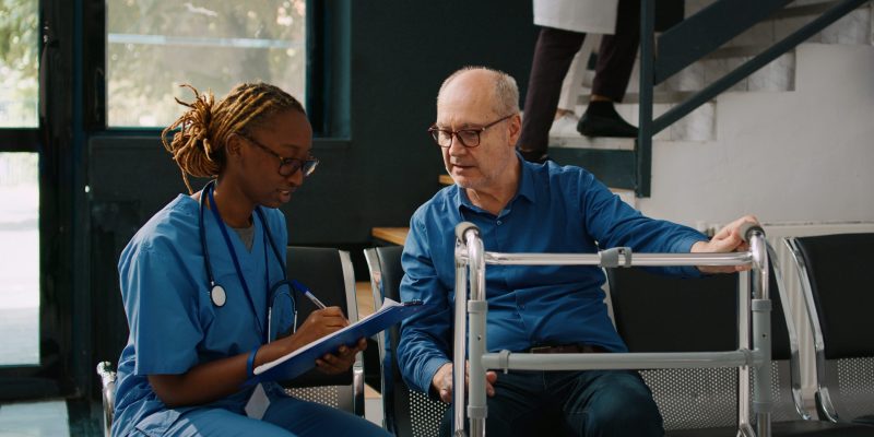 Female nurse doing checkup consultation with old patient in hospital waiting area, having walking frame. Medical assistant writing clinical report or appointment form, health center.